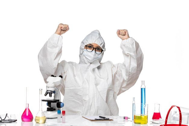 Vista frontal joven químico femenino en traje de protección especial sentado y regocijándose en el laboratorio de virus de escritorio blanco química covid