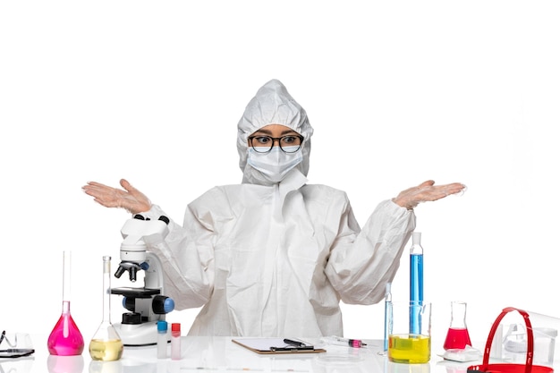 Vista frontal joven químico femenino en traje de protección especial sentado con soluciones en el laboratorio de virus de escritorio blanco química de salud covid