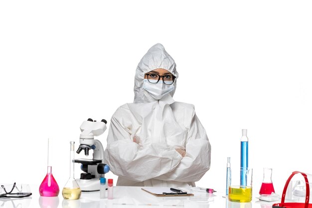 Vista frontal joven químico femenino en traje de protección especial sentado con soluciones sobre fondo blanco laboratorio de virus covid salud química