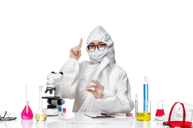 Vista frontal joven químico femenino en traje de protección especial sentado y trabajando sobre fondo blanco laboratorio de virus covid- química