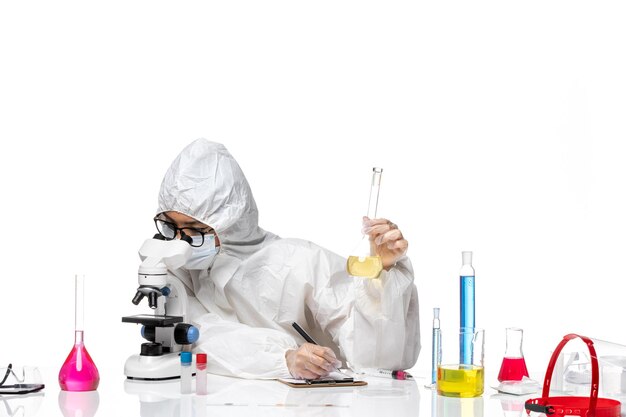 Vista frontal joven químico femenino en traje de protección especial sosteniendo el matraz con solución y usando microscopio sobre fondo blanco laboratorio química del virus covid