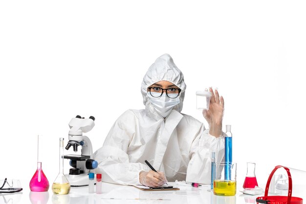 Vista frontal joven químico femenino en traje de protección especial sosteniendo el matraz vacío y escribiendo en el escritorio blanco laboratorio covid- salud del virus de la química