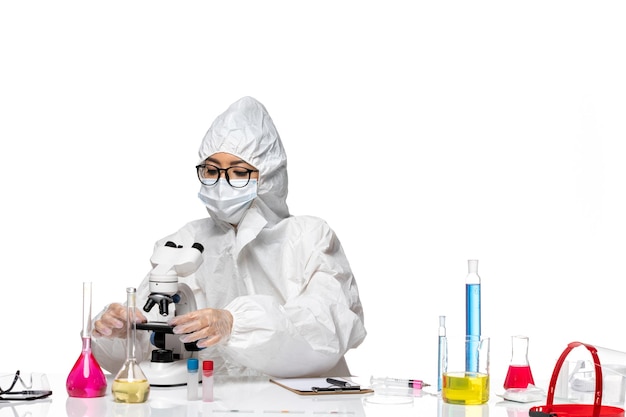 Vista frontal joven químico femenino en traje de protección especial trabajando con microscopio sobre fondo blanco covid- virus de laboratorio de química