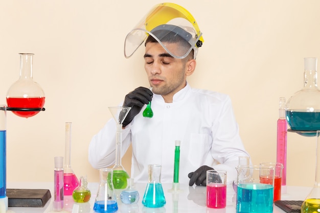 Vista frontal joven químico masculino en traje blanco especial frente a la mesa con soluciones de colores trabajando con ellos en el laboratorio de productos químicos de trabajo de ciencia de escritorio crema