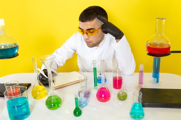 Vista frontal joven químico masculino en traje blanco frente a la mesa con soluciones coloreadas anotando notas en la mesa de luz química de laboratorio de trabajo de ciencia