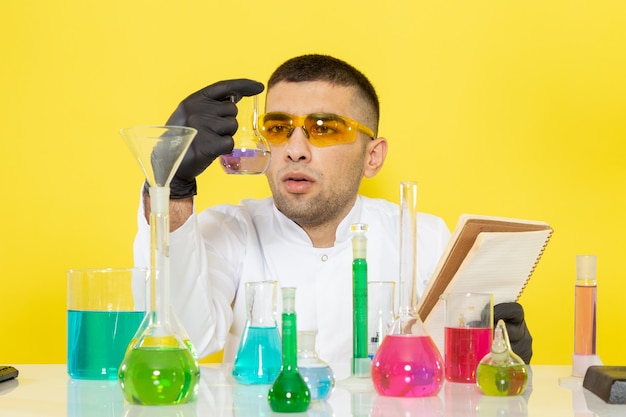 Vista frontal joven químico masculino en traje blanco frente a la mesa con soluciones coloreadas leyendo un bloc de notas en el escritorio amarillo ciencia trabajo química