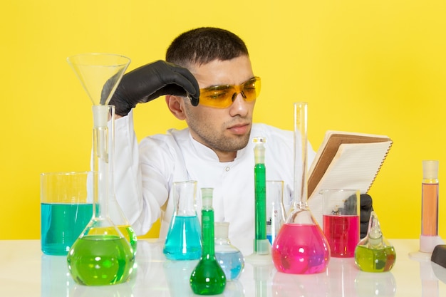 Vista frontal joven químico masculino en traje blanco frente a la mesa con soluciones coloreadas leyendo un bloc de notas en el escritorio amarillo trabajo de ciencia química de laboratorio