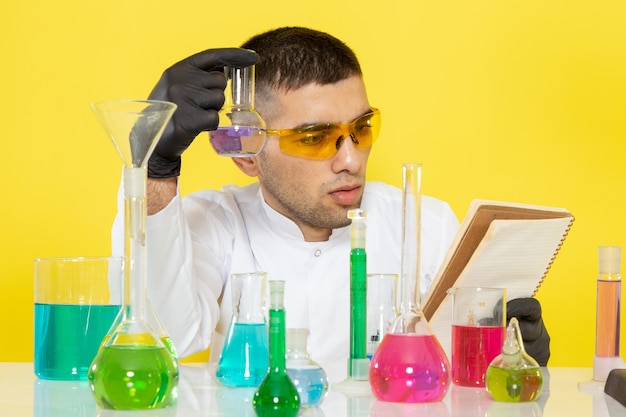 Foto gratuita vista frontal joven químico masculino en traje blanco frente a la mesa con soluciones coloreadas leyendo un bloc de notas en el escritorio amarillo trabajo de ciencia química de laboratorio