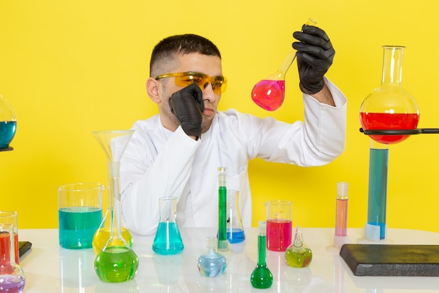 Vista frontal joven químico masculino en traje blanco frente a la mesa con soluciones coloreadas sosteniendo el matraz y pensando en el laboratorio de trabajo científico de escritorio amarillo