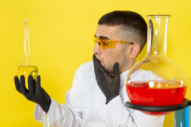 Vista frontal joven químico masculino en traje especial blanco frente a la mesa con soluciones coloreadas sosteniendo el matraz sorprendido en el escritorio amarillo trabajo científico laboratorio químico