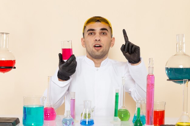 Vista frontal joven químico masculino en traje especial blanco sentado frente a la mesa con soluciones sosteniendo un matraz rosa y pensando en la ciencia química de laboratorio de pared crema