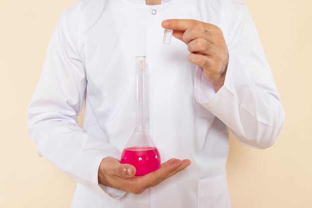 Vista frontal joven químico masculino en traje especial blanco sosteniendo frascos con solución rosa en la pared crema experimento de laboratorio de ciencias química científica