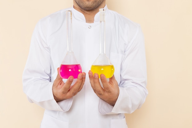 Vista frontal joven químico masculino en traje especial blanco sosteniendo frascos con soluciones amarillas y rosadas en la pared crema química experimento de laboratorio de ciencias