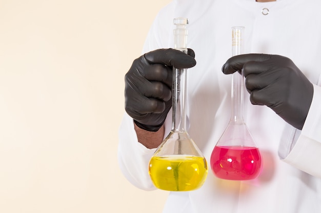 Foto gratuita vista frontal joven químico masculino en traje especial blanco sosteniendo pequeños frascos con soluciones en el experimento de ciencia química de laboratorio de pared crema