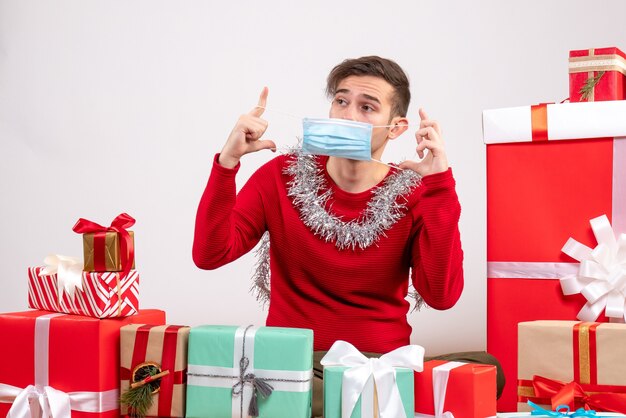 Vista frontal joven quitándose la máscara sentado alrededor de regalos de Navidad