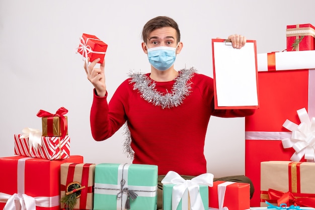 Vista frontal joven con regalo y portapapeles sentado alrededor de regalos de Navidad