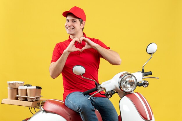 Vista frontal del joven romántico vestido con blusa roja y sombrero entregando pedidos haciendo gesto de corazón sobre fondo amarillo