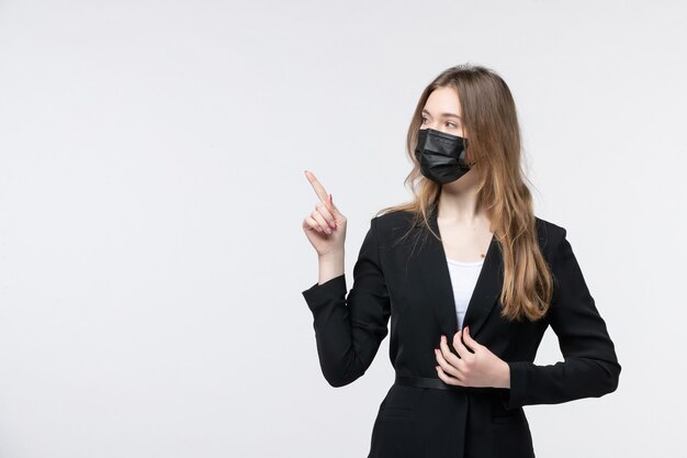 Vista frontal de la joven rubia en traje con mascarilla quirúrgica y apuntando hacia arriba en blanco