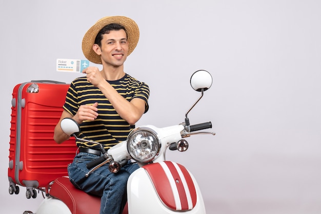 Vista frontal del joven satisfecho con sombrero de paja en ciclomotor con billete de avión