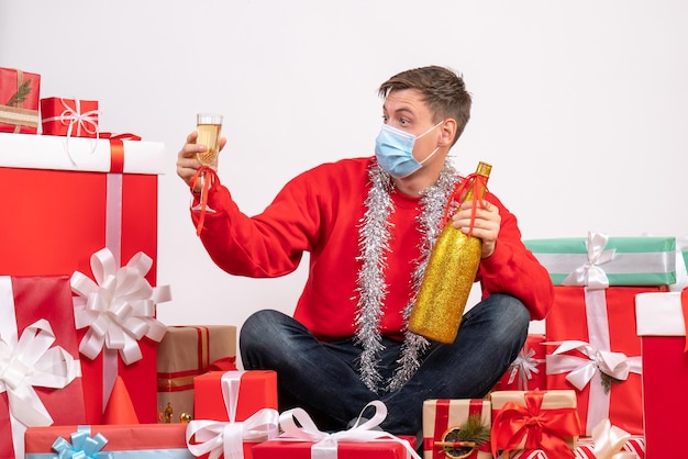 Vista frontal del joven sentado alrededor de regalos de Navidad celebrando con champán en la pared blanca