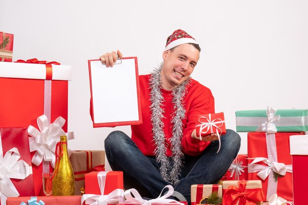 Vista frontal del joven sentado alrededor de regalos de Navidad con una nota en la pared blanca