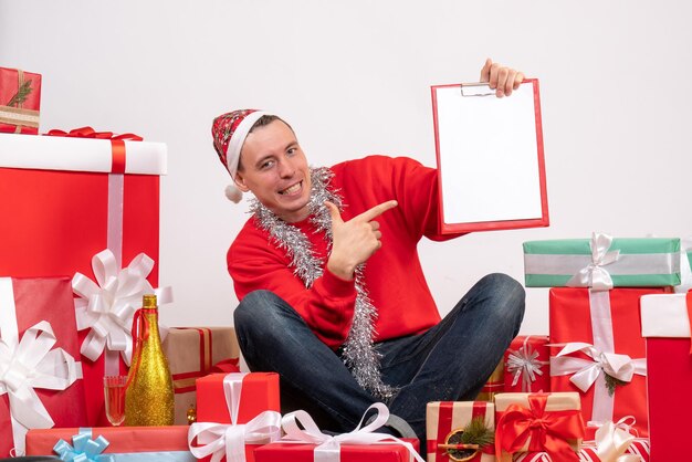 Vista frontal del joven sentado alrededor de regalos de Navidad con una nota en la pared blanca