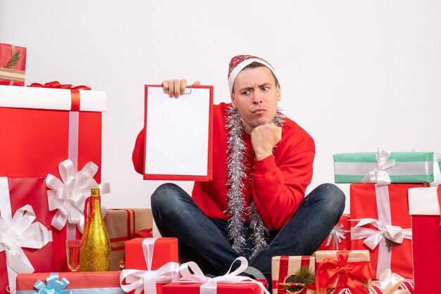 Vista frontal del joven sentado alrededor de regalos de Navidad con una nota en la pared blanca