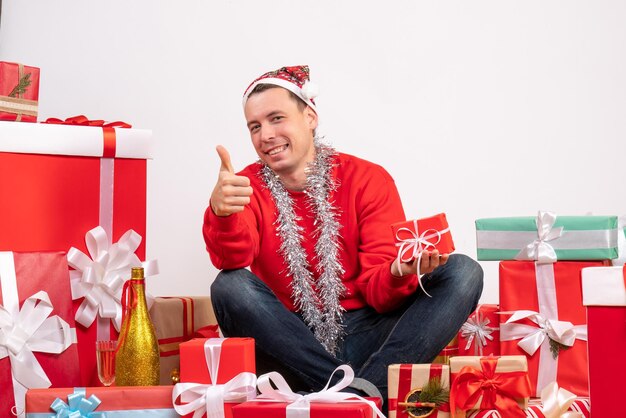 Vista frontal del joven sentado alrededor de regalos de Navidad en la pared blanca