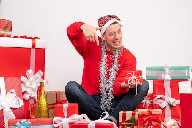 Vista frontal del joven sentado alrededor de regalos de Navidad en la pared blanca