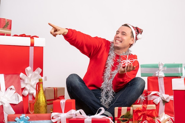 Vista frontal del joven sentado alrededor de regalos de Navidad en la pared blanca