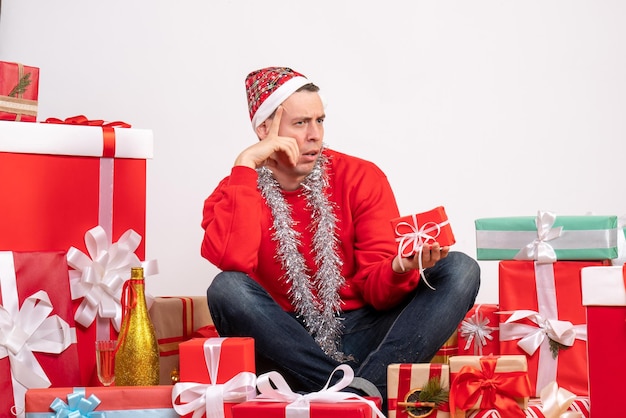 Vista frontal del joven sentado alrededor de regalos de Navidad en la pared blanca