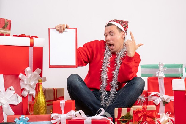 Vista frontal del joven sentado alrededor de regalos de Navidad con portapapeles en la pared blanca