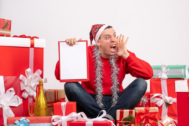 Vista frontal del joven sentado alrededor de regalos de Navidad con portapapeles en la pared blanca