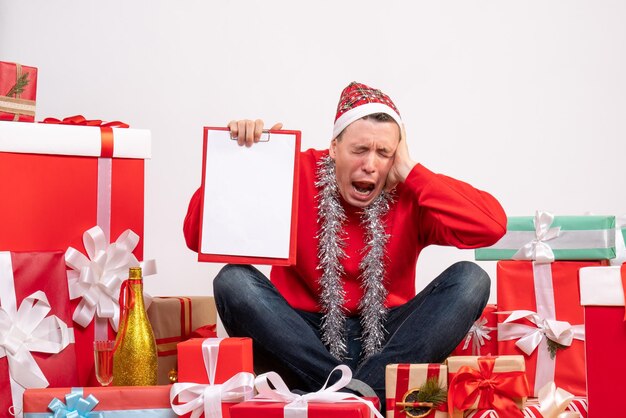 Vista frontal del joven sentado alrededor de regalos de Navidad con portapapeles en la pared blanca