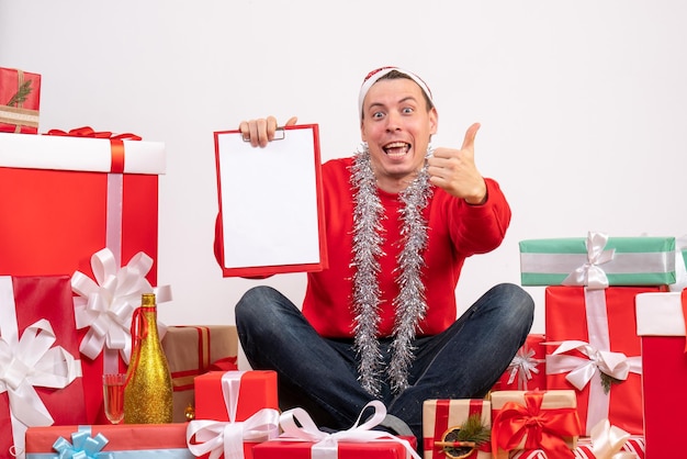 Vista frontal del joven sentado alrededor de regalos de Navidad con portapapeles en la pared blanca