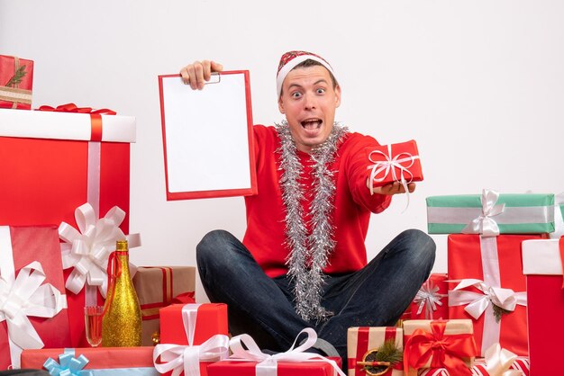 Vista frontal del joven sentado alrededor de regalos de Navidad con portapapeles en la pared blanca