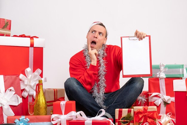 Vista frontal del joven sentado alrededor de regalos de Navidad con portapapeles en la pared blanca