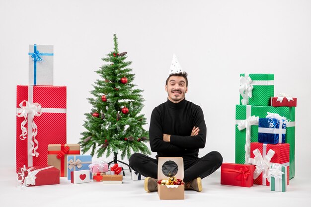 Vista frontal del joven sentado alrededor de regalos navideños y sonriendo en la pared blanca