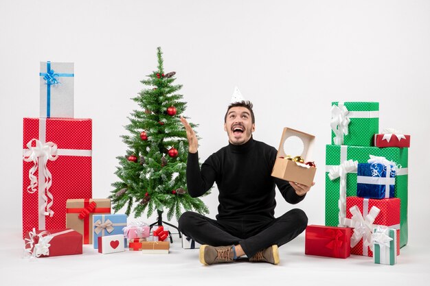 Vista frontal del joven sentado alrededor de regalos navideños sosteniendo juguetes de árbol en la pared blanca