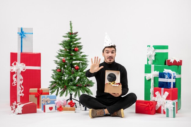 Vista frontal del joven sentado alrededor de regalos navideños sosteniendo juguetes de árbol en la pared blanca