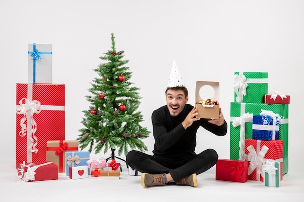 Vista frontal del joven sentado alrededor de regalos navideños sosteniendo juguetes de árbol en la pared blanca