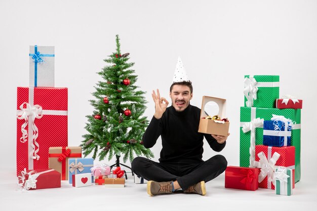 Vista frontal del joven sentado alrededor de regalos navideños sosteniendo juguetes de árbol en la pared blanca