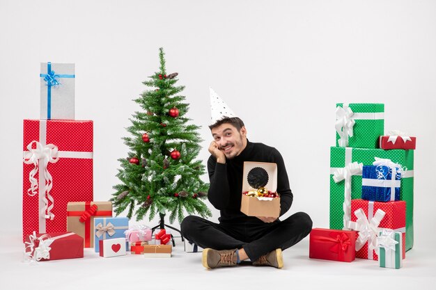 Vista frontal del joven sentado alrededor de regalos navideños sosteniendo juguetes de árbol en la pared blanca