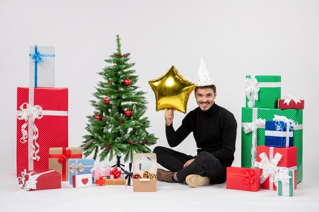 Vista frontal del joven sentado alrededor de regalos y sosteniendo la figura de una estrella dorada en la pared blanca