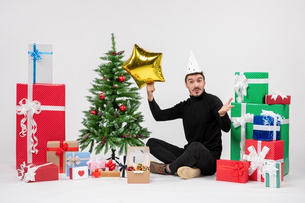 Vista frontal del joven sentado alrededor de regalos y sosteniendo la figura de una estrella dorada en la pared blanca