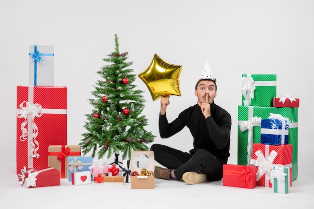 Vista frontal del joven sentado alrededor de regalos y sosteniendo la figura de una estrella dorada en la pared blanca