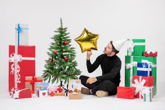 Vista frontal del joven sentado alrededor de regalos y sosteniendo la figura de una estrella dorada en la pared blanca