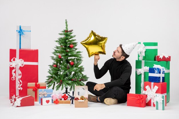 Vista frontal del joven sentado alrededor de regalos y sosteniendo la figura de una estrella dorada en la pared blanca