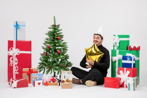 Vista frontal del joven sentado alrededor de regalos y sosteniendo la figura de una estrella dorada en la pared blanca