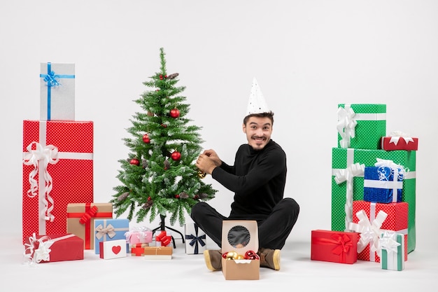 Vista frontal del joven sentado alrededor de regalos de vacaciones decorando arbolito en la pared blanca